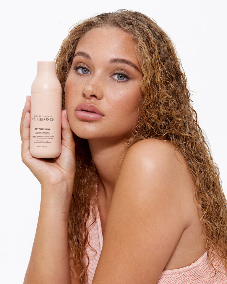 A orange, blonde model with long curly hair in a pink top, holding a bottle of leave-in treatment for the hair.
